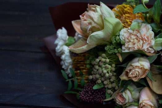 En bukett med blandade blommor och grönt lövverk ligger på ett mörkt bord.