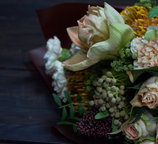 En bukett med blandade blommor och grönt lövverk ligger på ett mörkt bord.